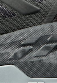 Schwarzer Sportschuh mit strukturiertem Obermaterial, das geometrische Muster aufweist, Gummisohle und ausgeprägten Rillenakzenten für Grip und Unterstützung.