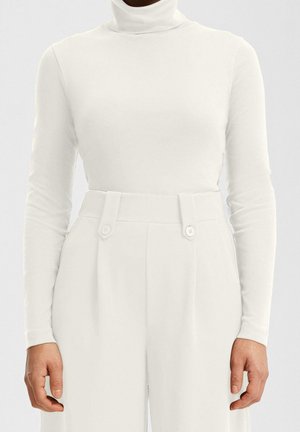 Personne portant un top à manches longues à col roulé blanc, rentré dans un pantalon blanc taille haute avec passants de ceinture boutonnés.