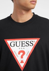 Musta puuvillainen t-paita, jossa on suuri punavalkoinen kolmionmuotoinen logo, keskellä teksti "GUESS U.S.A." ja kysymysmerkki.