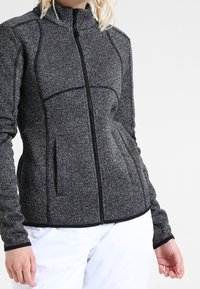 Woman wearing a fitted, zip-up gray textured jacket with black trim and white pants, standing against a plain light background.