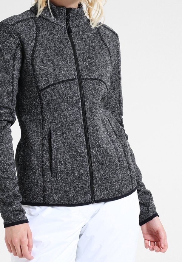 Mujer vestida con una chaqueta ajustada de textura gris con cremallera y ribetes negros, y pantalones blancos, de pie contra un fondo claro y liso.