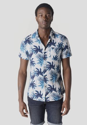 Joven con cabello corto y rizado que lleva una camisa blanca de manga corta con estampado de palmeras azules y pantalones cortos de mezclilla oscuros enrollados.