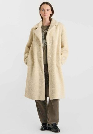 Roomkleurige, fluffy oversized jas met een ingegraven revers, knopen aan de voorkant en twee zijzakken. Draag het over een groene blouse en bruine broek.