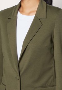 Blazer vert olive avec une coupe ajustée, col à revers, fermeture à un bouton et tissu texturé. Comprend une poche latérale et une doublure lisse.