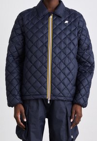 Veste matelassée bleu marine avec une fermeture éclair à l'avant, comportant une bande jaune, blanche et rouge le long de la fermeture éclair ainsi qu'un petit logo sur la poitrine.