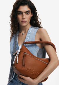 Borsa a tracolla in pelle marrone con superficie testurizzata, chiusura con zip e manico intrecciato. Presenta un nastro decorativo bianco e un logo impresso.
