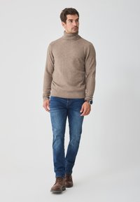 Barna bordás mandzsettával és szegéllyel rendelkező bézs garbópulóver, kék farmernadrággal és barna bőr csizmával párosítva, világos háttér előtt állva.