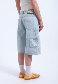 Pantaloni cargo in denim azzurro chiaro con molte tasche, orli rifiniti e vestibilità rilassata. Indossati con scarpe sportive gialle e rosa e calzini bianchi.