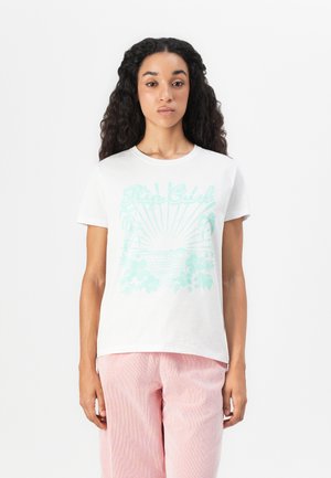 Woman with long curly hair wearing a white T-shirt with blue tropical print and pink pants standing against a plain white background.