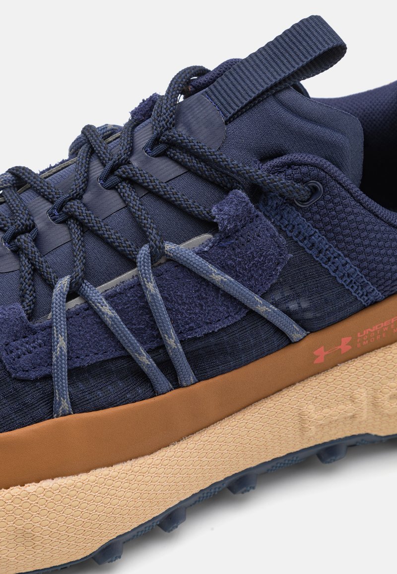 Męski but sportowy w kolorze granatowym, z teksturowanej tkaniny, kontrastującymi zamszowymi akcentami, jasnobrązową podeszwą środkową oraz grubą podeszwą z wypustkami zapewniającymi przyczepność.