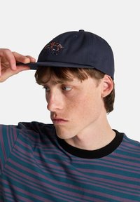 Μπλε ναυτικό καπέλο με κεντητό σχέδιο σε κόκκινο και πορτοκαλί. Το μοντέλο φοράει ριγέ μπλουζάκι σε τιρκουάζ και μωβ με μαύρο λαιμό.