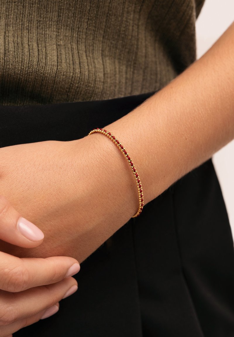 Pulsera dorada con acentos en rojo y verde, con un diseño delicado de pequeñas cuentas esféricas. Lleva puesta en una muñeca sobre un fondo oscuro.