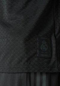 Černý sportovní dres s texturovaným vzorem. Obsahuje černou látkovou etiketu s logem Real Madrid a textem "EST. 1902." Jednoduchý design.