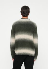 Homem de cabelo escuro curto, vestindo umsuéter felpudo listrado em verde e branco, mostrado por trás, contra um fundo branco simples.