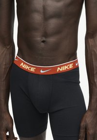 Αρσενικός κορμός που φοράει μαύρα boxer briefs με πορτοκαλί ζώνη μέσης που έχει το λογότυπο Nike και τα σύμβολα swoosh.