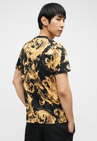 Νεαρός άνδρας με κοντά μαύρα μαλλιά, που φοράει μαύρο μπλουζάκι με χρυσά περίτεχνα μπαρόκ σχέδια, κοιτάζοντας πάνω από τον ώμο του.