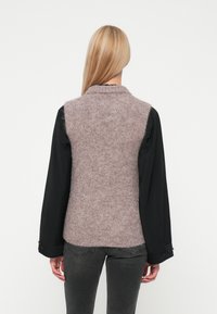 Pihe-puha rózsaszín-szürke gyapjú keverékből készült kötött mellény, galléros kialakítással, fekete hosszú ujjú inggel párosítva. Az alján szürke farmer látható.