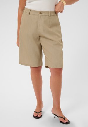 Kvinde iført beige knælange shorts med knap og lynlås, hvid top, sorte hælede sandaler og gyldt armbånd på ensfarvet baggrund.