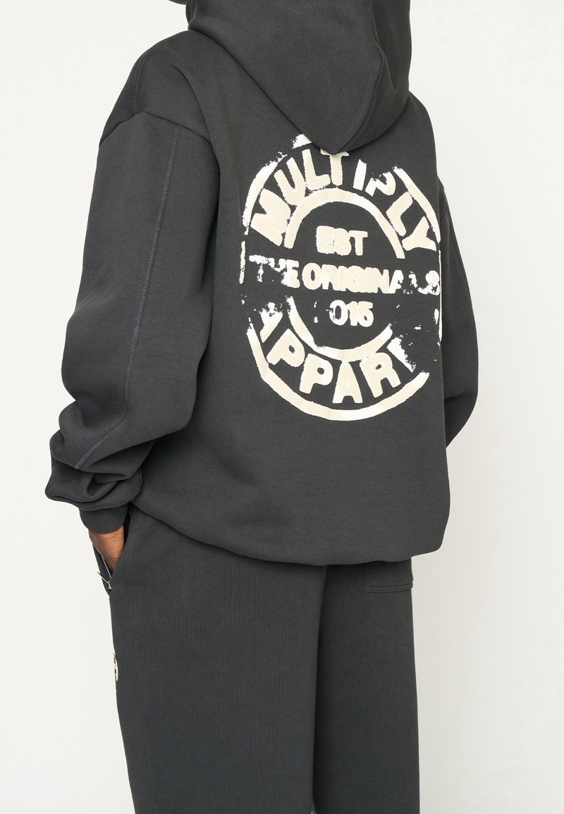 Un hoodie gris foncé avec un grand imprimé blanc usé portant l'inscription "MULTIPLY EST 2016 APPAREL" au dos. Il possède une capuche avec cordon de serrage et une coupe décontractée.