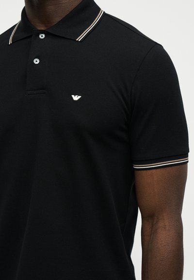 Μαύρο κοντομάνικο πόλο μπλουζάκι με λευκό και καφέ ριγέ γιακά και άκρες μανικιών, δύο κουμπιά και μικρό λευκό λογότυπο στο αριστερό στήθος.