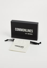 Čierna obdĺžniková krabička označená "COMMONLINES [ BY / BROTHERS ]" vedľa krémového vrecúška so šnúrkou, na ktorom je rovnaké logo v čiernej farbe.