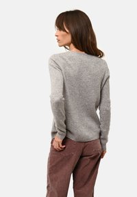 Žena s hnědými vlasy, oblečená v dlouhém světle šedém svetru s dlouhým rukávem a hnědých kalhotách, stojí zády k fotoaparátu na bílém pozadí.