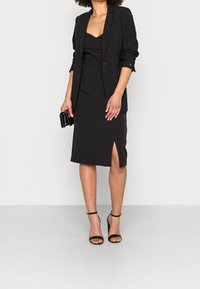Robe noire ajustée avec une fente, superposée sous un blazer noir. Le tissu semble lisse ; des sandales à talons hauts complètent le look. Aucun motif voyant.