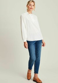 Weißes Blusenshirt mit Rüschenkragen und langen Ärmeln, kombiniert mit blauen Skinny Jeans und braunen Mokassins. Glatte Textur, lässiges Design.