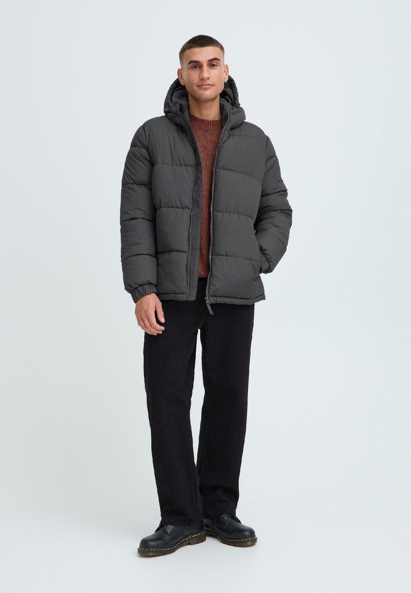 Mørkegrå pufferjakke med hætte, quiltet design, frontlynlås og sidelommer. Båret over en brun sweater og mørke bukser.