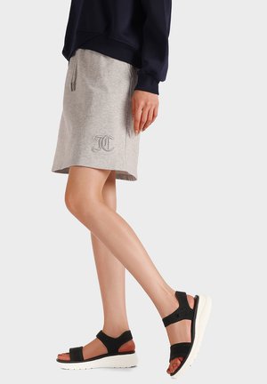 Persona che indossa una gonna grigia al ginocchio con logo ricamato e sandali neri con suole bianche platform, in piedi davanti a uno sfondo semplice.