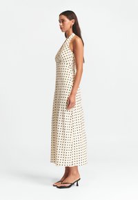 Vestido maxi de lunares en beige claro con puntos negros, que presenta un escote halter y una silueta fluida. Combinado con sandalias de tacón negras.