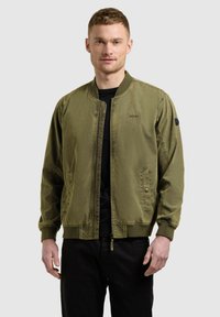 Homme portant une veste bomber vert olive sur une chemise noire et un pantalon noir, debout devant un fond clair uni.