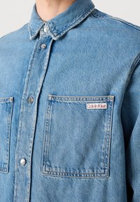 Světle modrá džínová košile s klasickým límečkem, dvěma předními kapsami a štítkem s logem Calvin Klein. Disponuje viditelným prošíváním a detaily s knoflíky.