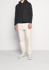 Černá péřová vesta přes námořnickou mikinu, doplněná krémovými kalhotami a béžovými vysokými teniskami. Vícevrstvý design s texturovanou látkou.