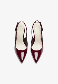 Décolleté slingback in pelle lucida bordeaux con punta affusolata, lati intagliati e una sottile cinghia alla caviglia. Texture liscia, branding minimo visibile.