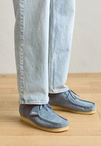 Blauwe suède mocassinschoenen met een lichtbruine rubberen zool, voorzien van strikveters en een klein label aan de zijkant, gecombineerd met lichtblauwe spijkerbroeken.