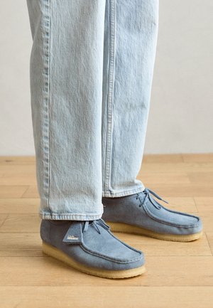 Blå ruskinds mokkasin sko med en lysebrun gummisål, der har sløjfebånd og et lille mærke på siden, parret med lyse denimjeans.