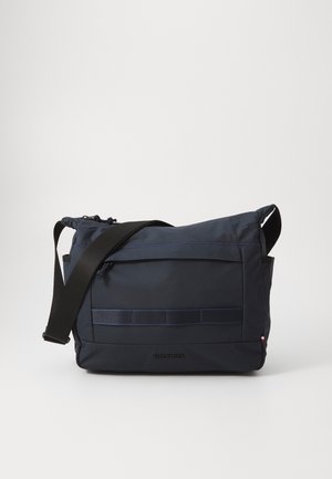 Ciemnoniebieska torba typu crossbody z regulowanym czarnym paskiem, przednią kieszenią na zamek oraz subtelnym logo na dole, na środku.