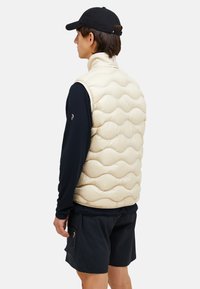 Beige Puffer-Weste mit wellenförmigem Steppmuster, hohem Kragen und leichtem Material, getragen über einem dunklen Langarmhemd und kombiniert mit schwarzen Shorts.