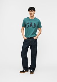 Νεαρός άνδρας που φοράει τιρκουάζ μπλούζα GAP, σκούρα τζιν με φαρδιά πόδια και μαύρα αθλητικά παπούτσια με λευκά κορδόνια, στέκεται με τα χέρια στις τσέπες.