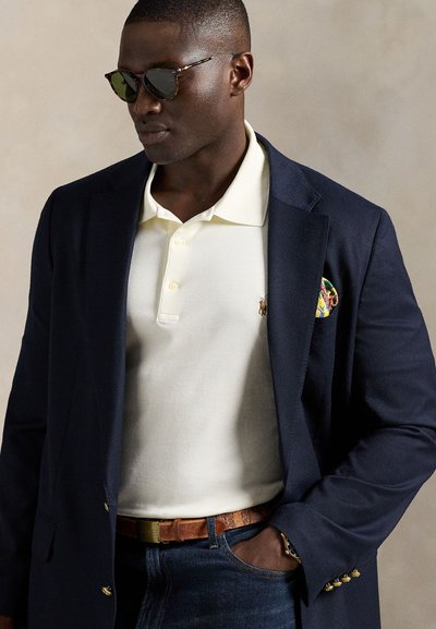 Marineblå blazer, kremfarget poloshirt med krage, brunt skinnbelte og mønstret lommetørkle; solbriller gir et moderne preg.