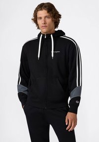 Felpa nera con zip, strisce bianche sulle spalle, dettagli grigi sulle maniche, tasche frontali e logo sul petto. Tessuto morbido e strutturato.