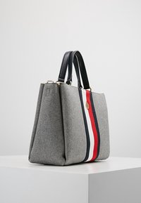 Grå håndtaske med marineblå læderhåndtag, med et centralt rødt og marineblå stribe og guld hardwaredetalje. Struktureret design med vinklet form.