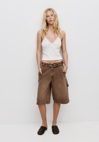 Bruna denim Bermuda-shorts med avslappnad passform, fickor och ett nitarprytt brunt bälte. Matchat med en vit linne med spetsdetaljer och mörka slip-on skor.