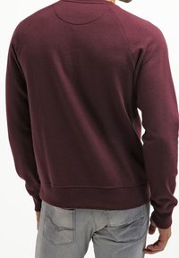 Felpa color bordeaux con scollo rotondo e maniche raglan, caratterizzata da un tessuto strutturato e polsini e orlo a costine.