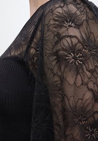 Μαύρο ύφασμα δαντέλας με λουλούδια, ανοιγμένο πάνω σε ριμπ δερμάτινο ρούχο, δείχνοντας περίπλοκα διαφανή λουλουδένια σχέδια και αντίθεση υφής στην περιοχή του ώμου.