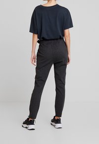 Osoba stojící zády, oblečená v tmavě šedých elastických kalhotách, černobílých teniskách a volném černém tričku na jednobarevném pozadí.