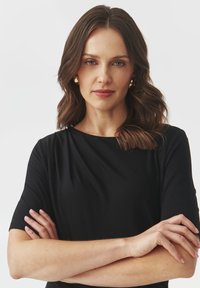Juodas trumpomis rankovėmis suknelė su apvalia iškirpte, lygiu paviršiumi ir prigludusia siluetu. Modelis demonstruoja sukryžiuotas rankas ir subtilius makiažo akcentus.