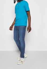 Camiseta de algodón azul con logo en el pecho, combinada con jeans ajustados azul oscuro y zapatillas beige con acentos multicolores.