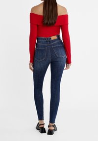 Jeans ajustados de tiro alto en azul oscuro con un diseño de corte en la parte trasera, que cuentan con dos bolsillos traseros y un dobladillo deshilachado. Combinados con una blusa roja de escote bardot.